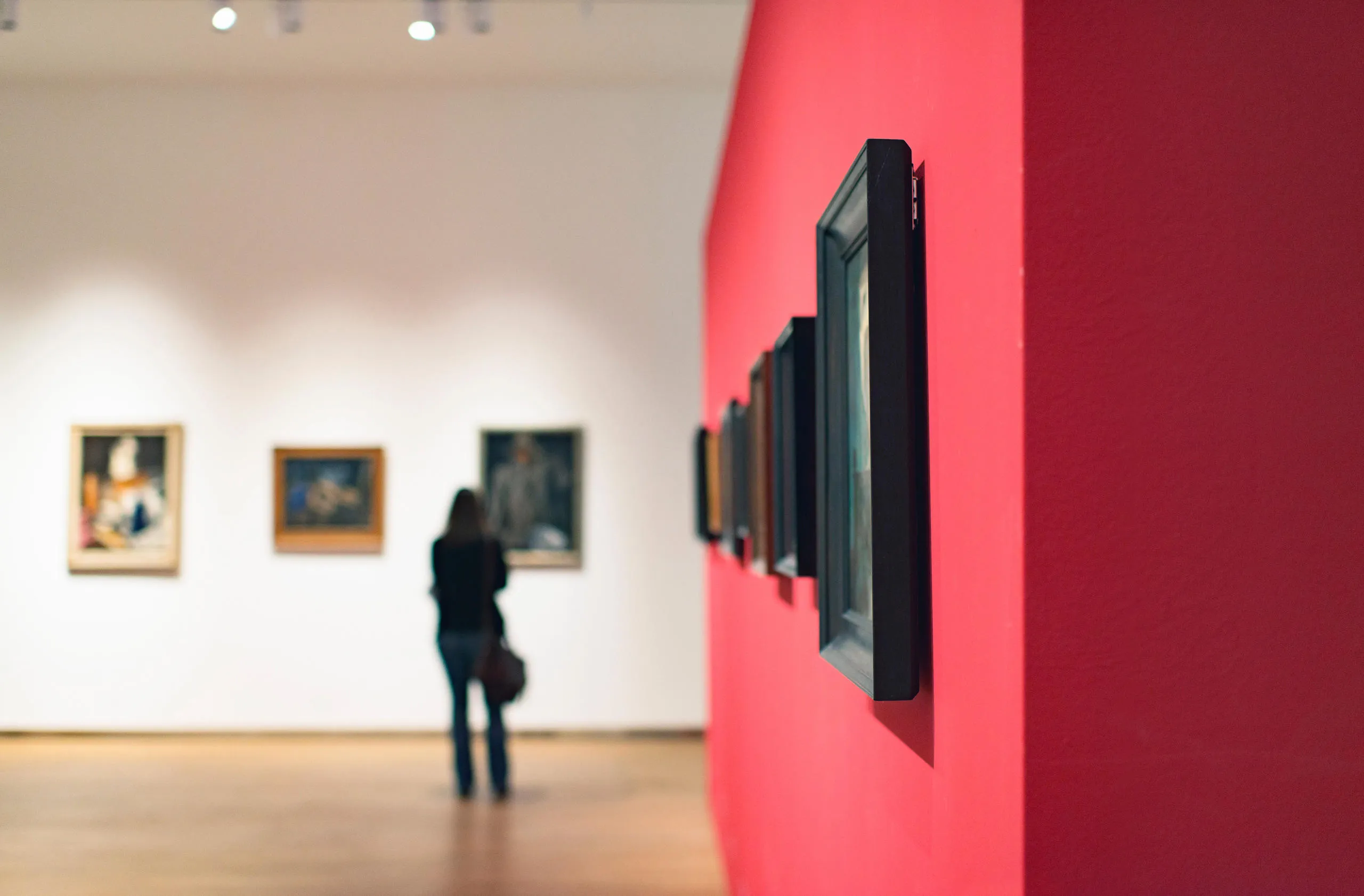 Gallery space with a red wall with artworks and a person in the distance